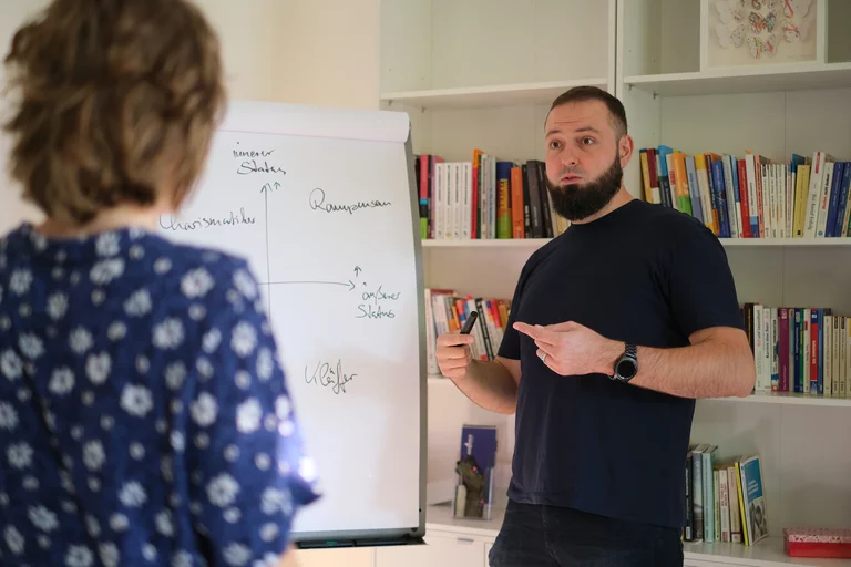 Coachingsituation mit einer Flipchart. Dr. Sebastian Hümbert-Schnurr erläutert eine Skizze, eine Klientin steht ihm zugewandt im Vordergrund.